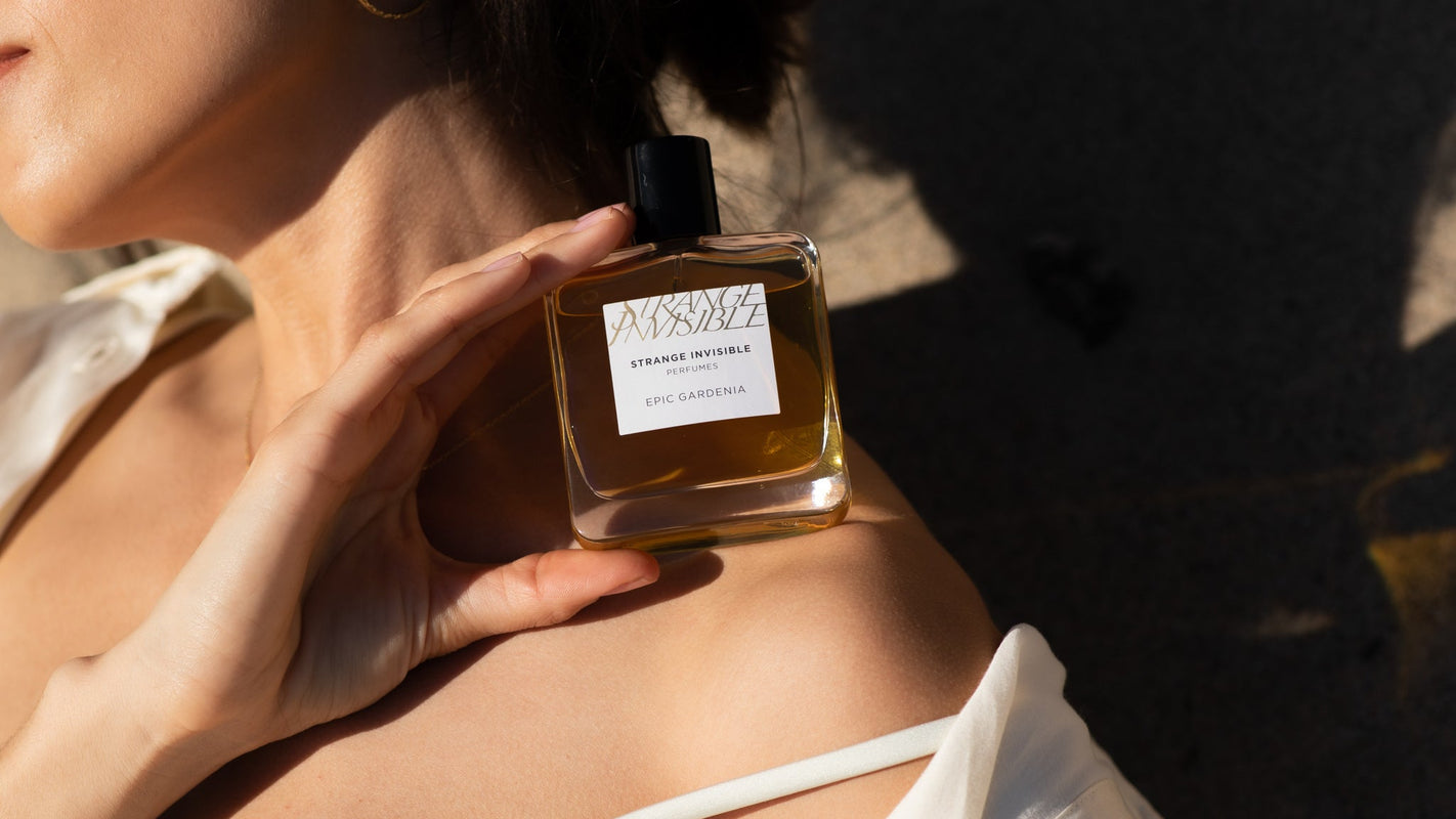 A woman in a white top holds a square glass perfume bottle labeled 'Epic Gardenia' against warm sunlight.